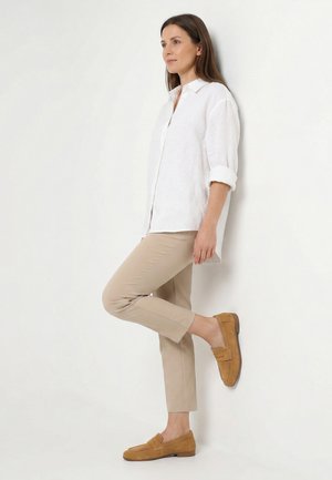 Vrouw in beige cropped broek, wit shirt met lange mouwen en opgerolde manchetten, en beige loafers, staand met één gebogen been tegen een witte muur.