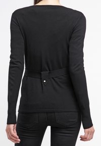 Kvinna iförd en figurnära svart långärmad topp med ett tygbälte och svarta byxor, vänd bort från kameran mot en vit bakgrund.