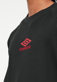Pogled od blizu na osebo, ki nosi črno dukserico z rdečim izvezenim logotipom Umbro na prsih, na beli podlagi.