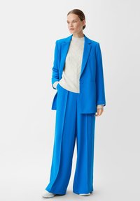 Ein hellblaues maßgeschneidertes Blazer, kombiniert mit einer passenden weiten Hose. Darunter ein cremafarbener Strickpullover mit Rautenmuster und gerippten Bündchen.