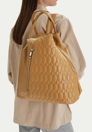 Žena v béžové bundě nese velký hnědý prošívaný kožený batoh se zlatým zipem a logem značky na boku.