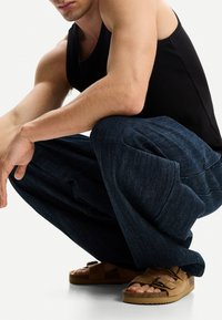 Temno modre široke kavbojke z teksturirano površino in visokim pasom, združene z rjavimi semišnimi natikači z dvojno zanko.