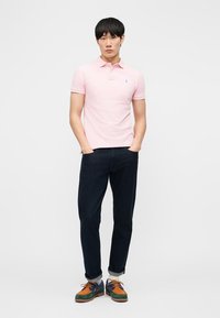 Vyras stovintis su rankomis kišenėse, dėvintis šviesiai rausvą polo marškinėlį, tamsias susuktas kelnes ir margaspalvius laisvalaikio batus ant balto fono.