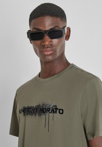 Camiseta verde oliva con un logo pintado en negro con spray. El modelo lleva unas gafas de sol rectangulares negras, que muestran un acabado suave y mate.