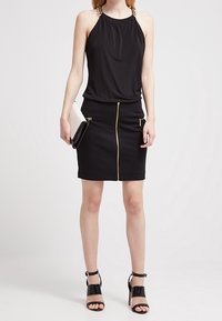 Top noir à encolure halter avec un décolleté froncé ; jupe crayon avec une fermeture éclair frontale et des accents dorés ; accompagnée de sandales à talons noires et d'une pochette.