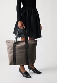 Liela pelēka auduma iepirkumu soma ar melnu siksnu, uz kuras ir iespiests zīmola nosaukums. Soma ir ar elegantu, vienkāršu dizainu un to tur cilvēks.