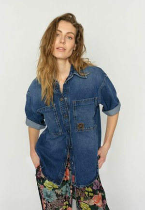 Naine, kellel on pikad lainelised juuksed, kannab sinist teksasärki, mille varrukad on rullitud, ja värvilisi lillemustriga pükse, seisab käed taskutes.