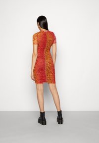 Vestido bodycon com mangas curtas, em degradê de laranja e vermelho, padrão floral, com detalhes franzidos nas laterais, usado com botas negras até o tornozelo.