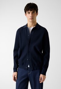 Cardigan lavorato a maglia blu navy con scollo con colletto, bottoni frontali e polsini a coste, abbinato a una T-shirt bianca e jeans di denim scuro.