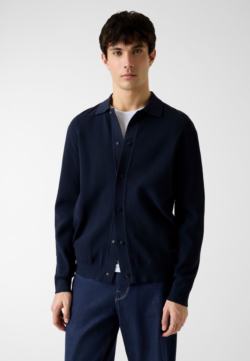 Cardigan lavorato a maglia blu navy con scollo con colletto, bottoni frontali e polsini a coste, abbinato a una T-shirt bianca e jeans di denim scuro.