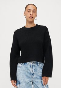 Zwarte gebreide truien met een gestructureerde afwerking, voorzien van een ronde hals en losvallende lange mouwen, gecombineerd met lichtblauwe versleten jeans.