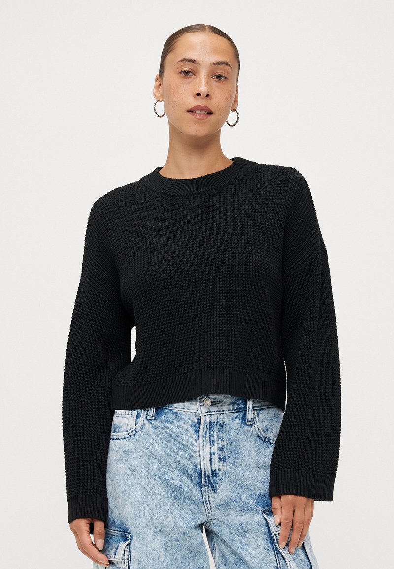 Zwarte gebreide truien met een gestructureerde afwerking, voorzien van een ronde hals en losvallende lange mouwen, gecombineerd met lichtblauwe versleten jeans.