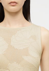 Gros plan d'une personne portant un haut sans manches beige avec un motif floral texturé et un design en maille.