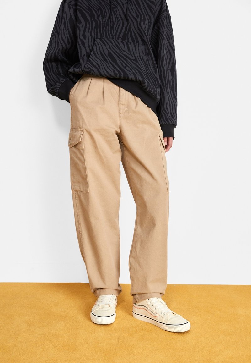 Personne portant un pantalon cargo beige avec des poches latérales, des baskets blanches et un sweat à capuche noir à motifs zébrés, debout sur un sol moutarde.