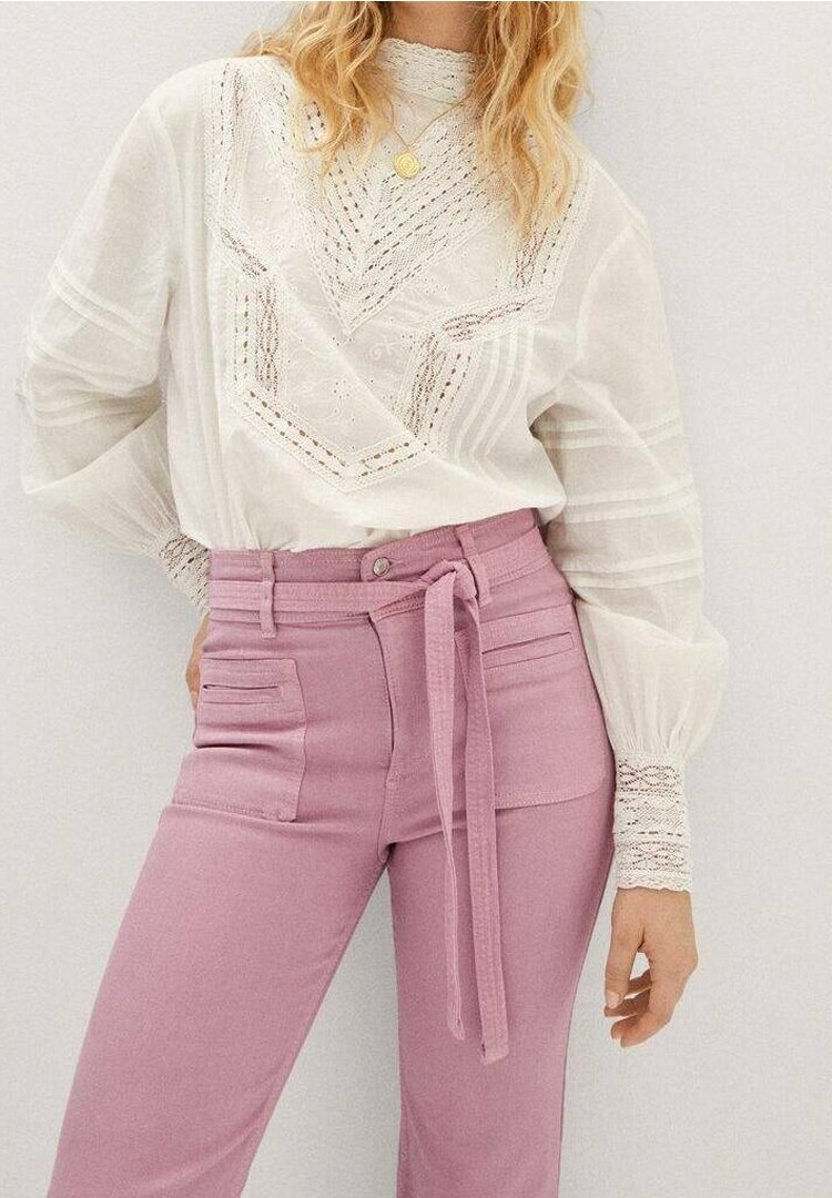 Femme portant un chemisier blanc en dentelle à manches longues, rentré dans un pantalon rose taille haute avec une ceinture à nouer et des poches plaquées à l'avant.
