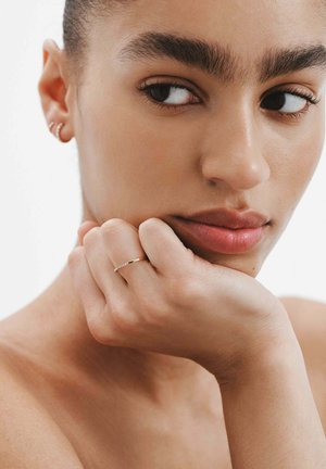 Mujer joven con cejas gruesas descansando su barbilla en la mano, lleva un anillo delgado y aretes pequeños en forma de aro, mirando hacia un lado.