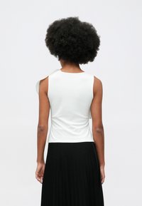 Person with curly hair wearing a sleeveless white top and a black pleated skirt, facing away against a plain light background.