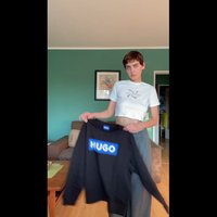 Μαύρη φούτερ μπλούζα με μπλε λογότυπο "NUGO", με χαλαρή εφαρμογή και ριμπ μανσέτες. Το μοντέλο φοράει ένα άσπρο κοντό τοπ και γκρι παντελόνι.