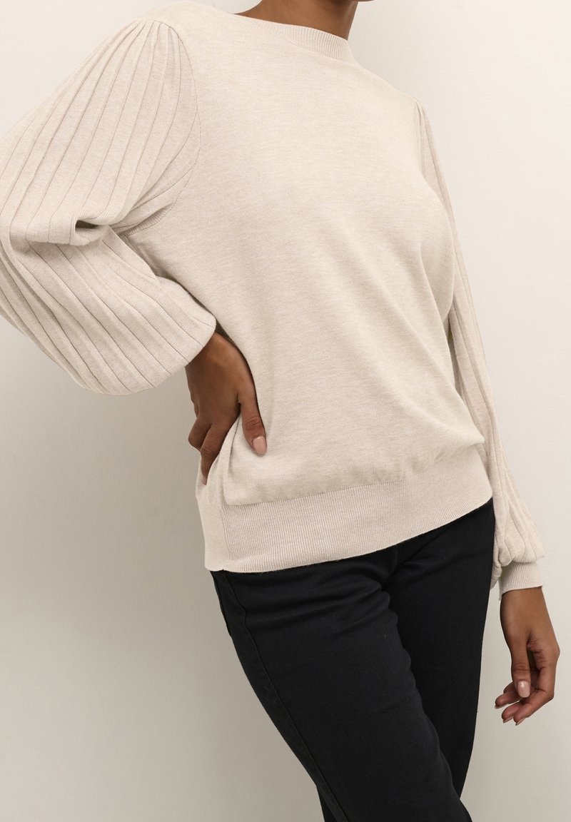 Femme portant un pull côtelé beige avec des manches bouffantes et un pantalon noir, posant avec une main sur la hanche.