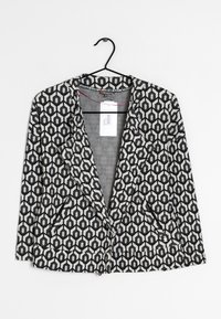 Svart och vit mönstrad kavaj med hexagonal design, med sjalkrage och två framfickor, hängande på en svart galge.