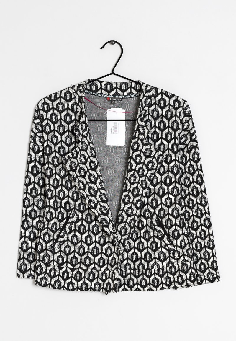 Svart och vit mönstrad kavaj med hexagonal design, med sjalkrage och två framfickor, hängande på en svart galge.