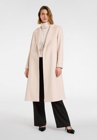 Jasnoróżowy długi płaszcz z wyłogowym kołnierzem, dwoma bocznymi kieszeniami i dopasowanym krojem, noszony na beżowym golfie i czarnych spodniach o szerokich nogawkach.