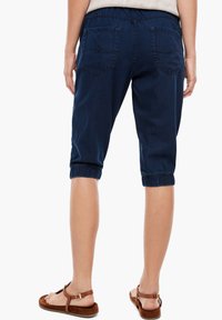 Denimcaprbyxor i mörkblått med elastiska muddremmar, med två bakfickor och en avslappnad passform. Bärs med bruna sandaler.