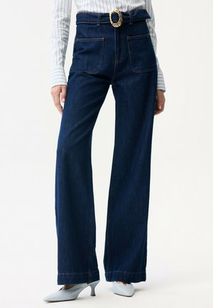 Jeans a gamba larga blu scuro a vita alta con grandi tasche frontali e cintura con fibbia rotonda dorata, indossati con décolleté azzurri a punta.