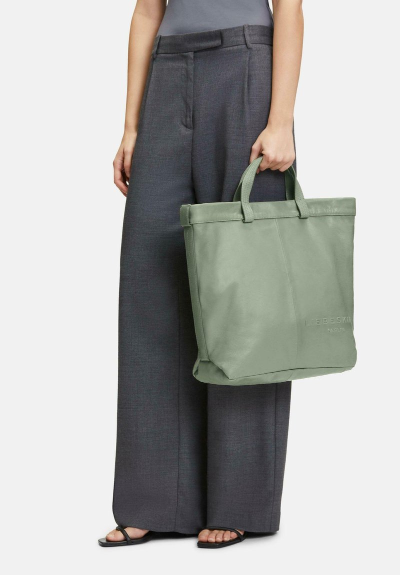 Sac fourre-tout en cuir vert avec poignées supérieures doubles, texture lisse et logo en relief subtil, associé à un pantalon gris ample.