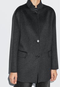 Mörkgrå ullblandad kappa med hög krage, knappslutning framtill och två sidofickor. Rakt snitt och textilstruktur.