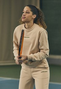 Felpa beige e pantaloni della tuta abbinati con il logo "BJÖRN BORG", che tiene in mano una racchetta da tennis arancione, su un campo indoor blu.