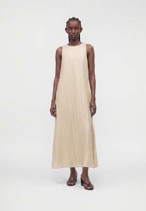 Personne grande avec des cheveux tressés portant une robe beige plissée sans manches, longueur cheville, et des sandales compensées marron, debout sur un fond uni.
