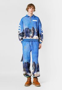 Conjunto de sudadera y joggers en azul, con un estampado de montañas. La sudadera tiene un cuello con cremallera y un logo blanco. Botas en color marrón con suela gruesa.