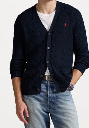 Mann trägt eine dunkelblaue Zopfstrick-Cardigan über einem weißen T-Shirt, hellblaue Jeans und eine blaue Armbanduhr, mit einer Hand in der Tasche.