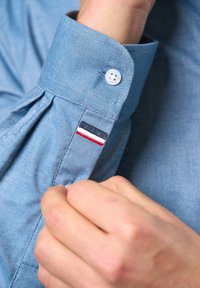 Blue chambray shirt cuff with a button, featuring a small embroidered label with red, white, and navy stripes, and pleated design details.