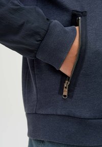 Navy zip-up jacket featuring a textured, ribbed hem and cuffs, with a side pocket highlighted by a silver zipper and smooth fabric accents.