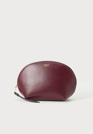 Καλλυντική θήκη από δέρμα μπορντό Lancel Paris με χρυστό φερμουάρ και λουράκι καρπού σε απλό ανοιχτόχρωμο φόντο.