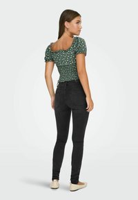 Grøn blomstret bluse med puffærmer og smocket krop, parret med højtaljede sorte jeans og beige flade sko, set bagfra.