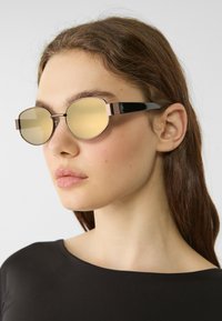 Kulaté sluneční brýle se zlatým nádechem, černými acetátovými nožičkami a kovovým rámem. Mají elegantní design s minimalistickou estetikou a ostrými akcenty.