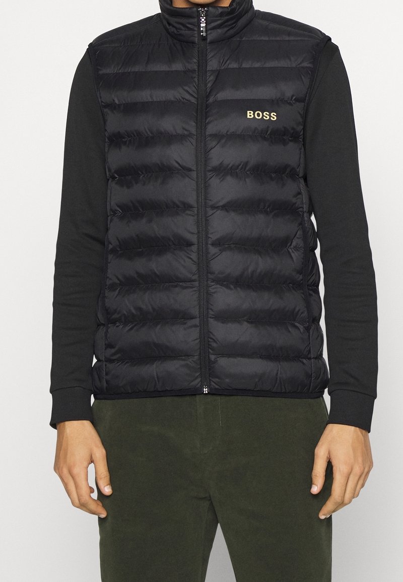 Man i svart vadderad ärmlös väst med "BOSS"-logotyp på bröstet, svart långärmad skjorta och mörkgröna byxor, som står rak.