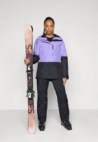 Μωβ και μαύρο μπουφάν για σκι, παντελόνι που ταιριάζει, κρατώντας ένα σκι με δέστρες, σε απλό φόντο. Υφή υφάσματος, λειτουργικός σχεδιασμός.