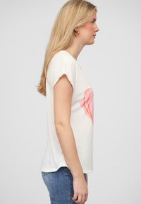 Ľahké biele bavlnené tričko s ružovým srdcom obkresleným v oranžovej farbe. Krátke rukávy a voľný strih. Nosené s modrými džínsami.