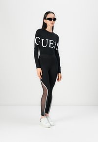 Crna majica dugih rukava s "GUESS" logotipom, uparen s crnim tajicama koje imaju prozirne panele i bijele bočne akcenta. Bijele tenisice upotpunjuju izgled.