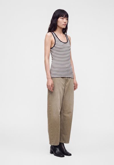 Femme aux cheveux noirs portant un débardeur rayé noir et blanc, un pantalon beige ample et des bottines noires, debout devant un fond uni.