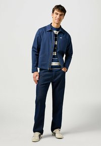 Chaqueta azul marino con cremallera y cuello puntiagudo, llevada sobre una camisa a rayas. Combinada con jeans de azul oscuro y zapatillas ligeras.