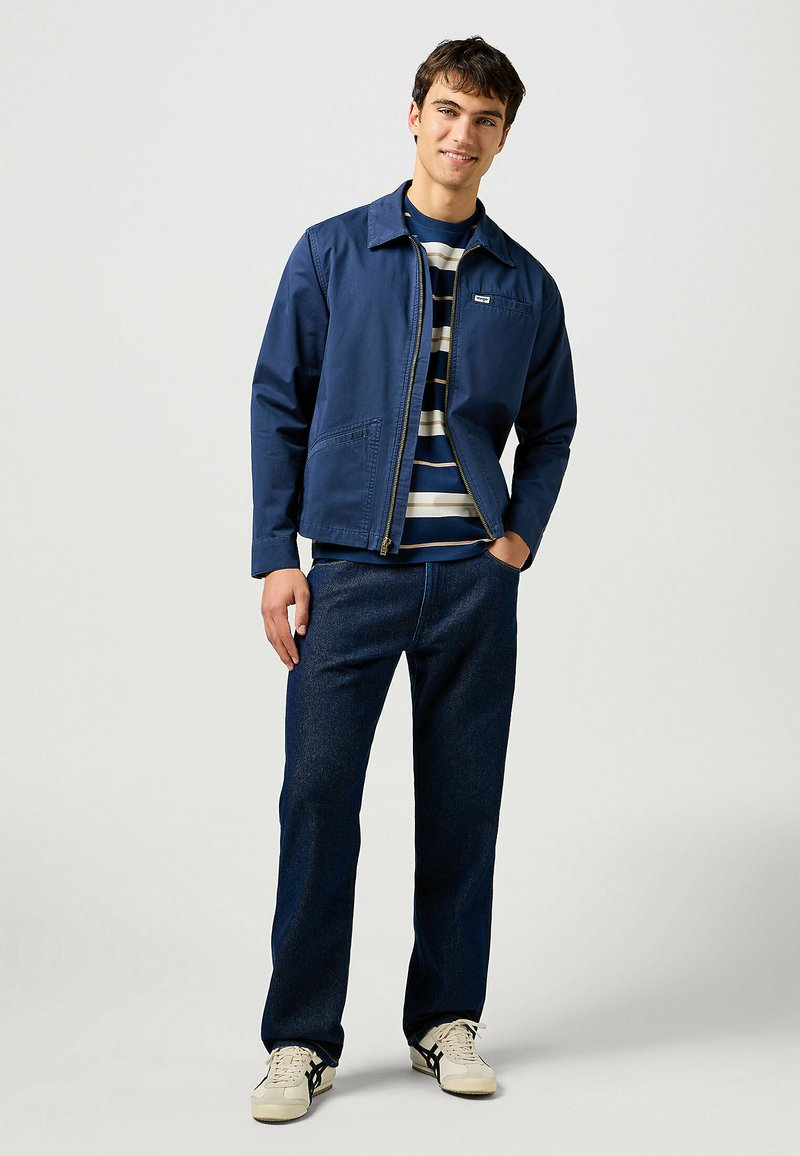 Granatowa kurtka zapinana na zamek błyskawiczny z wyciąganym kołnierzem, noszona na pasiakowej koszuli. W zestawieniu z ciemnoniebieskimi dżinsami i jasnymi sneakersami.