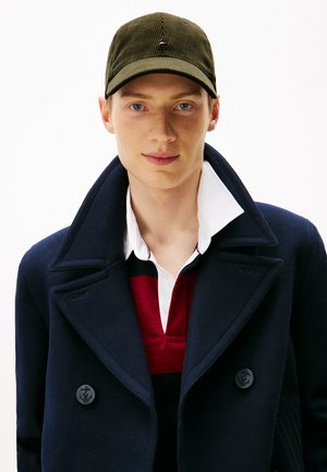 Young man with light skin and blue eyes wearing an olive green cap, navy blue double-breasted coat, and red, white, and black collared shirt.