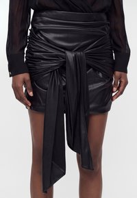 Mini-jupe noire en simili cuir avec des détails froncés et une longue ceinture nouée sur le devant, portée avec un chemisier noir transparent à manches longues.