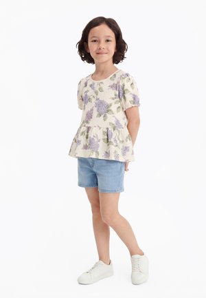 Rövid sötét hajú lány, aki virágmintás rövid ujjú blúzt, farmer rövidnadrágot és fehér tornacipőt visel, kezét a háta mögött tartva áll.