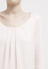 Helles rosa Bluse mit weitem Ausschnitt und langen Ärmeln, aus weichem, fließendem Stoff und vertikalen Falten nahe dem Ausschnitt.